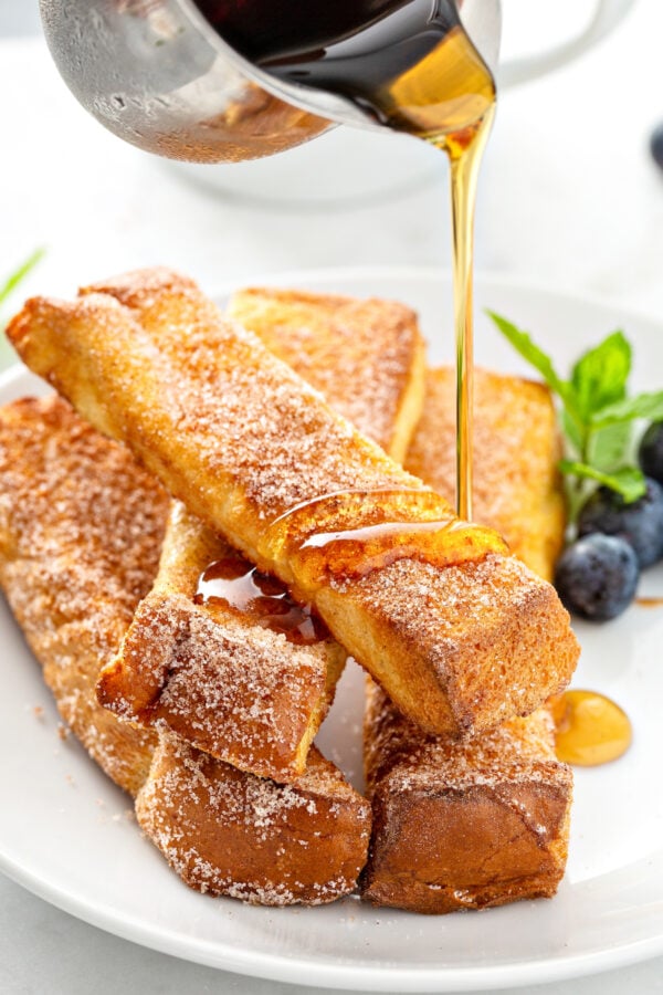 syrup is being poured over a stack of French toast sticks on a white plate.