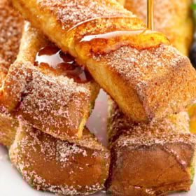 Close-up of golden brown Air Fryer French Toast sticks stacked on a plate, sprinkled with sugar and cinnamon, as syrup is poured over them.