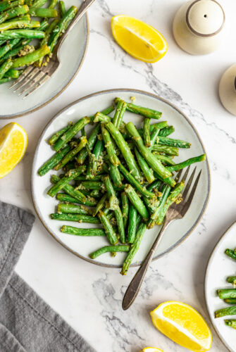 Air fryer green beans on a dinner plate with a fork. Lemon wedges and other plates of green beans are arranged artistically on the table.