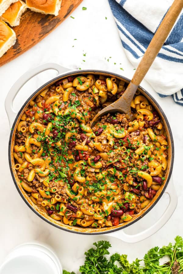 This easy, one-pot Beef Goulash recipe is comfort food at its best with cheesy ground beef and tender noodles in a robust tomato sauce. A cheesy, beefy skillet of goulash is being stirred with a large wooden spoon.