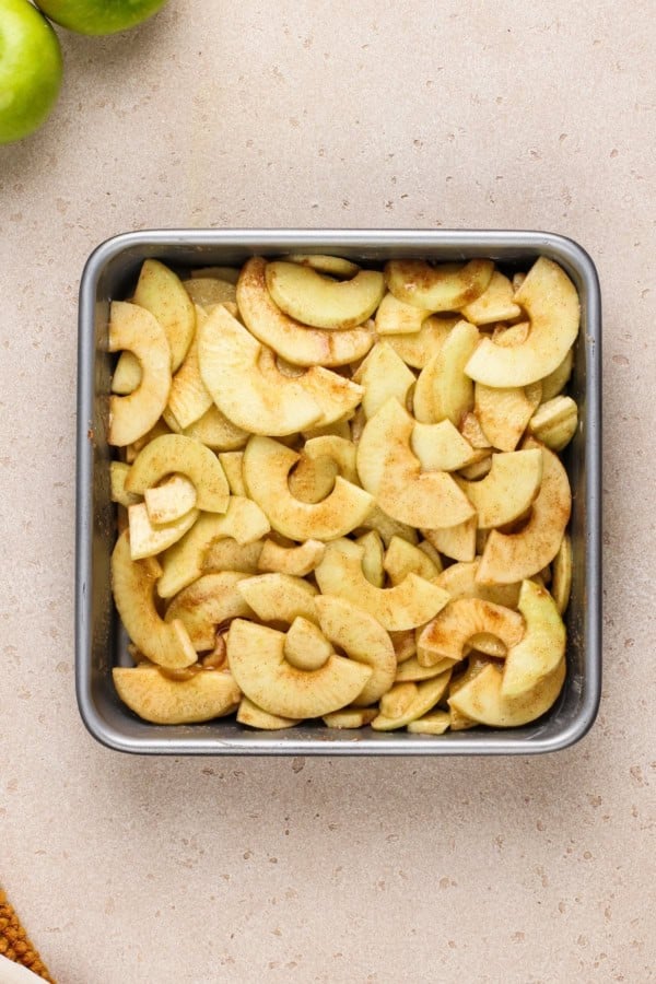 Old‑fashioned Apple Crisp is made with tender, juicy apples baked under a cinnamon‑oat crumble topping. Serve it warm with ice cream! A metal square baking pan filled with sliced apples coated in cinnamon and sugar sits on a light-colored countertop. Green apples are partially visible in the top left corner.