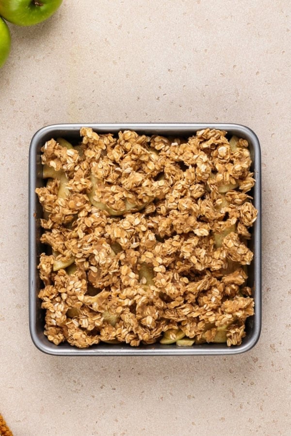 Old‑fashioned Apple Crisp is made with tender, juicy apples baked under a cinnamon‑oat crumble topping. Serve it warm with ice cream! A square baking dish filled with unbaked fruit crisp, topped with an oat and brown sugar crumble, sits on a light-colored countertop. Green apples are partially visible in the top left corner.