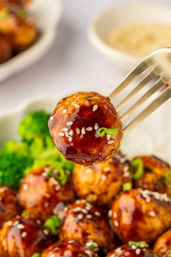 These sweet and savory Asian Chicken Meatballs are infused with fresh ginger and garlic, then baked and coated in a sticky honey-soy glaze. An asian chicken meatball is being lifted from a plate with a fork.
