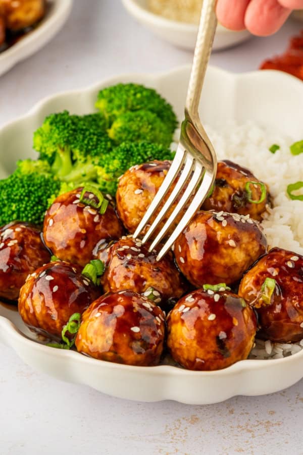 These sweet and savory Asian Chicken Meatballs are infused with fresh ginger and garlic, then baked and coated in a sticky honey-soy glaze. A fork is piercing a meatball on a plate.