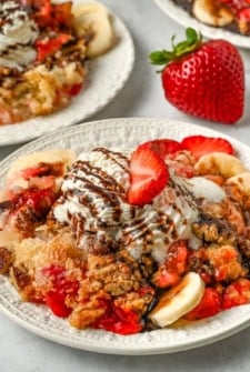 A plate of warm banana split cake topped with with strawberry and banana slices, crumbly topping, whipped cream, chocolate drizzle, and a strawberry slice on top.