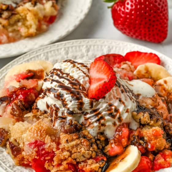 A plate of warm banana split cake topped with with strawberry and banana slices, crumbly topping, whipped cream, chocolate drizzle, and a strawberry slice on top.