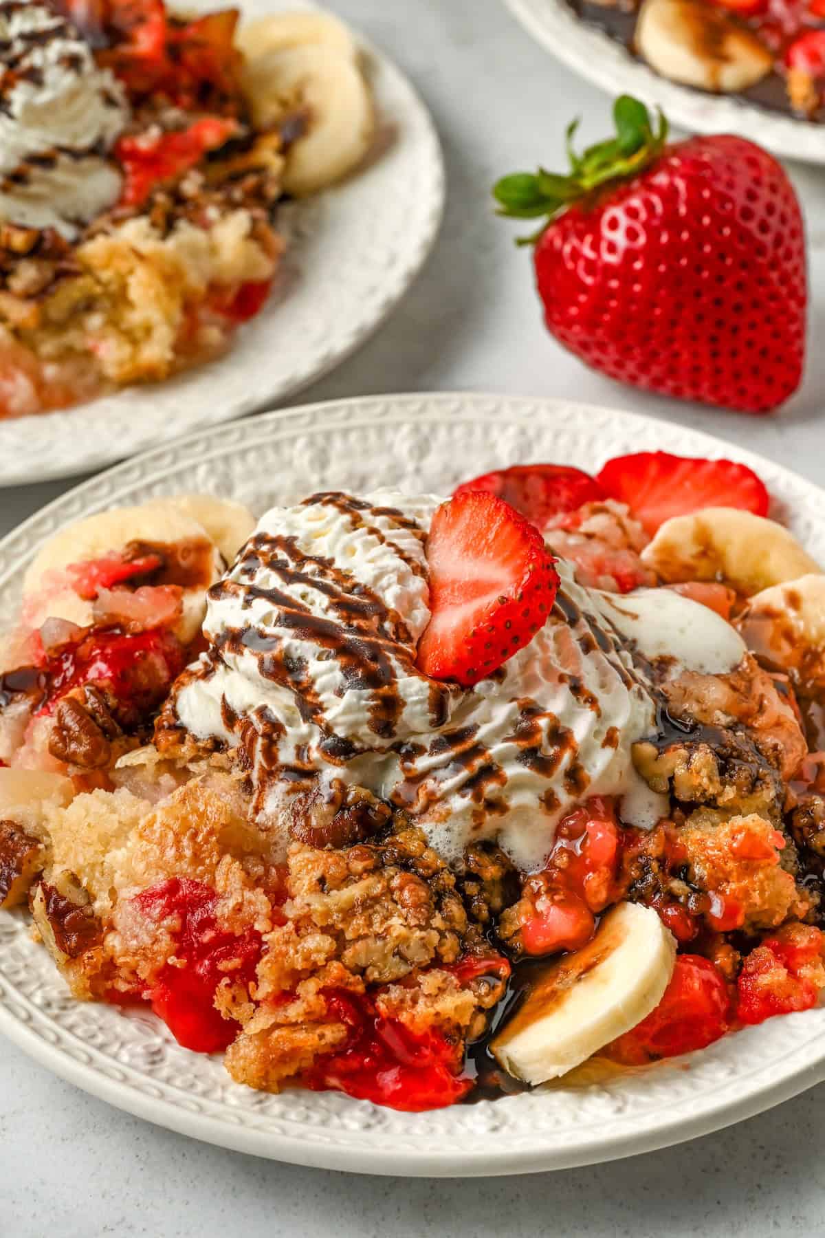 Full of pineapple, strawberries, bananas, and nuts, this easy Banana Split Dump Cake is served warm with whipped cream and chocolate syrup! A plate of warm banana split cake topped with with strawberry and banana slices, crumbly topping, whipped cream, chocolate drizzle, and a strawberry slice on top.