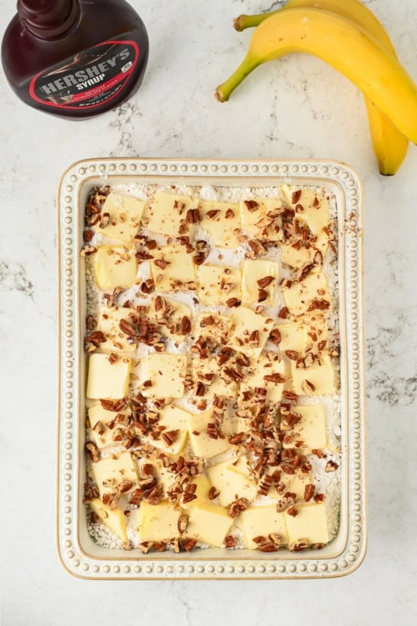 Full of pineapple, strawberries, bananas, and nuts, this easy Banana Split Dump Cake is served warm with whipped cream and chocolate syrup! Rectangular baking dish filled with cubed butter, chopped pecans, and other ingredients. A bottle of Hershey’s syrup and two bananas are nearby on a marble countertop.