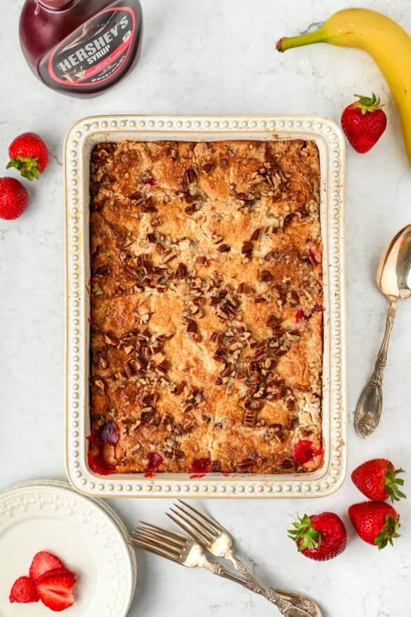 Full of pineapple, strawberries, bananas, and nuts, this easy Banana Split Dump Cake is served warm with whipped cream and chocolate syrup! A baked banana split cake topped with chopped pecans in a rectangular dish, surrounded by strawberries, a banana, a bottle of Hershey’s syrup, plates, forks, and a spoon on a white marble surface.