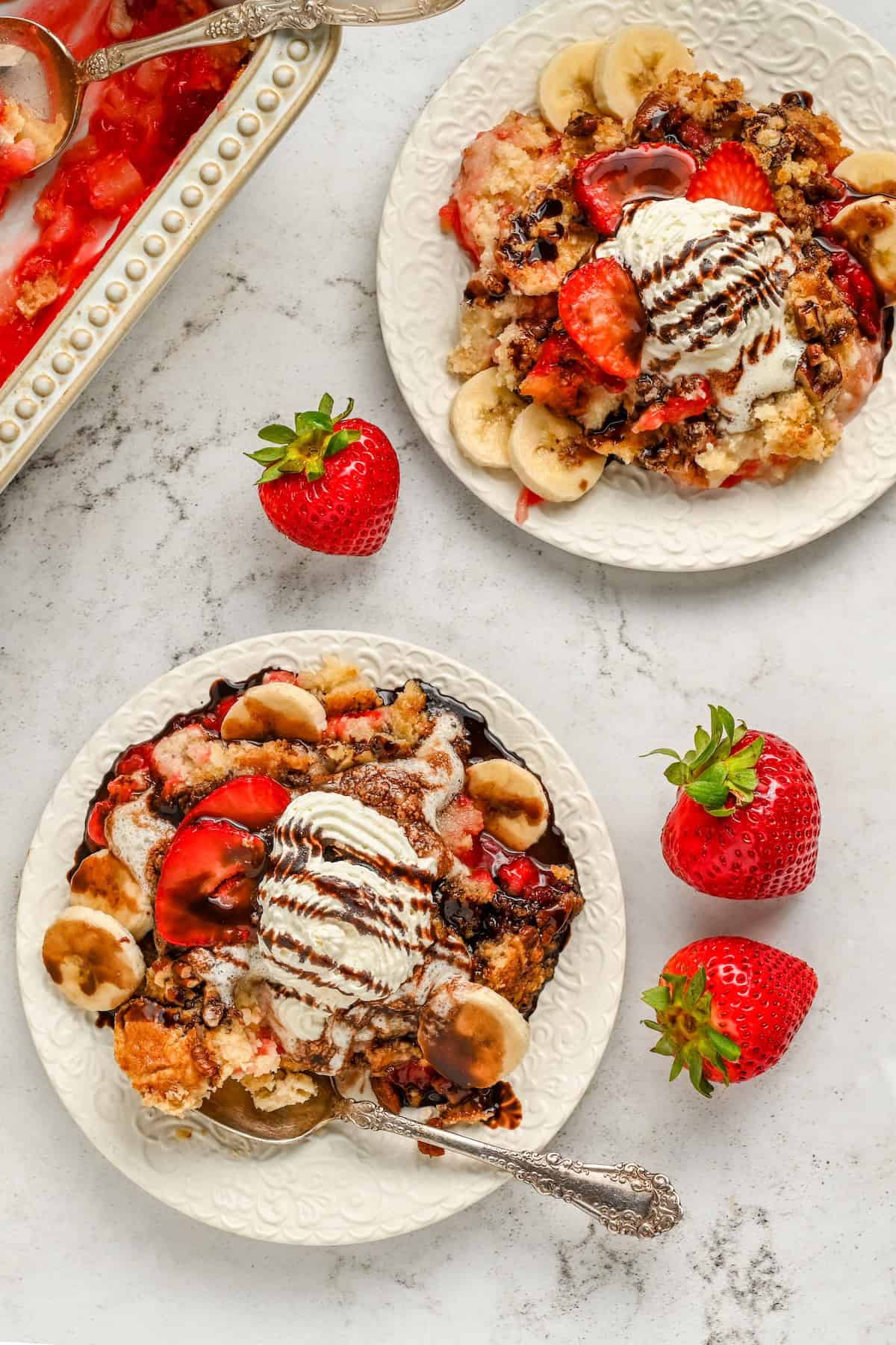Full of pineapple, strawberries, bananas, and nuts, this easy Banana Split Dump Cake is served warm with whipped cream and chocolate syrup! Two plates of banana split cake topped with sliced bananas, strawberries, vanilla ice cream, and chocolate sauce sit on a white surface, surrounded by fresh strawberries and a baking dish of the same dessert.