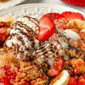 A plate of banana split dump cake topped with a scoop of vanilla ice cream, chocolate syrup, and a fresh strawberry slice, with bananas and cherries visible around the dessert.