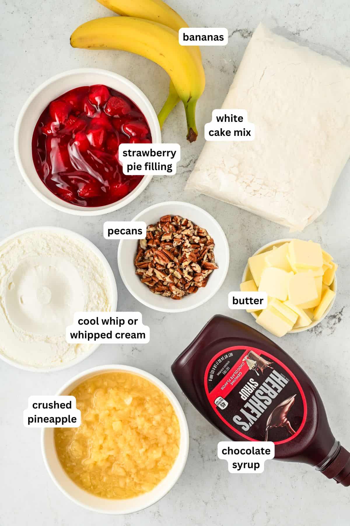 Full of pineapple, strawberries, bananas, and nuts, this easy Banana Split Dump Cake is served warm with whipped cream and chocolate syrup! Labeled ingredients for making banana split dump cake recipe on a countertop: bananas, white cake mix, strawberry pie filling, cool whip, crushed pineapple, chopped pecans, butter slices, and a bottle of chocolate syrup.
