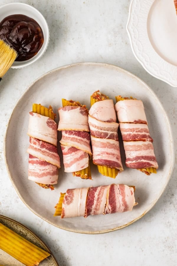 This mouthwatering bacon-wrapped BBQ Shotgun Shells recipe is the perfect smoky, cheesy, and meaty appetizer your guests will devour! Five uncooked manicotti shells stuffed and wrapped in bacon are neatly arranged on a white plate. A small bowl of bbq sauce with a brush lies nearby, set on a marble surface with additional plates visible at the edges.