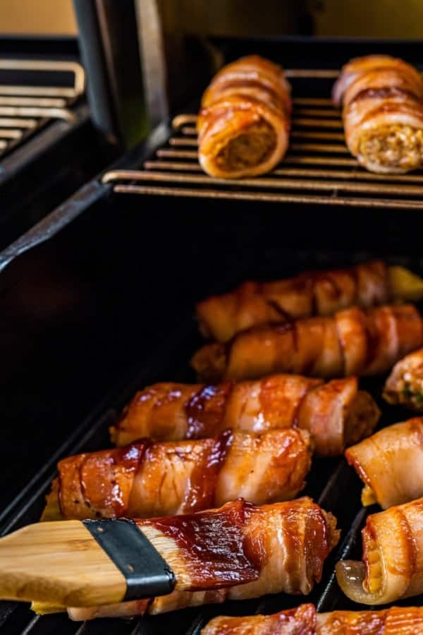 This mouthwatering bacon-wrapped BBQ Shotgun Shells recipe is the perfect smoky, cheesy, and meaty appetizer your guests will devour! Close-up of bacon-wrapped shotgun shells on a grill, brushed with a bbq sauce. More wrapped shells are visible on a higher rack inside the grill. The bacon looks crispy and browned.