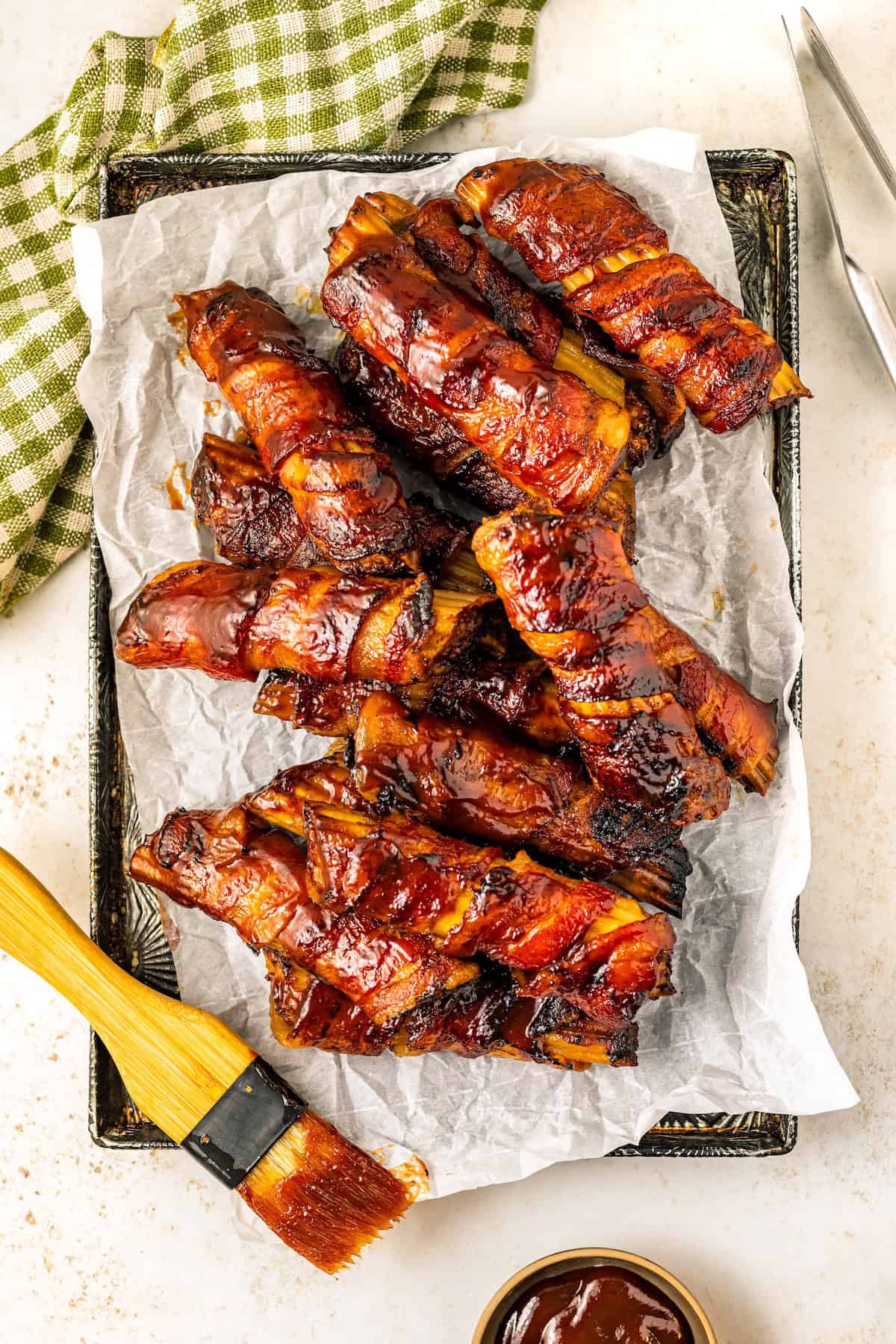 This mouthwatering bacon-wrapped BBQ Shotgun Shells recipe is the perfect smoky, cheesy, and meaty appetizer your guests will devour! Bacon wrapped BBQ shotgun shells are covered in bbq sauce and arranged on a parchment-lined tray. A basting brush with sauce is beside the ribs, and a small bowl of barbecue sauce is nearby. A green checkered cloth is in the upper left corner.