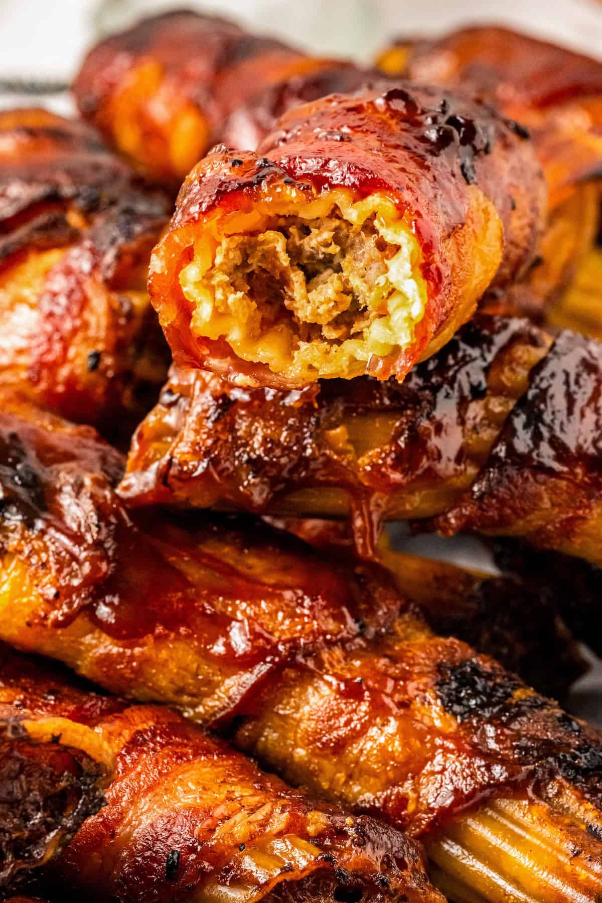 This mouthwatering bacon-wrapped BBQ Shotgun Shells recipe is the perfect smoky, cheesy, and meaty appetizer your guests will devour! Close-up of grilled, bbq sauce glazed meat-stuffed manicotti shells wrapped in bacon with a bite taken out of one to show the meaty interior. The exterior is crispy and caramelized, with a shiny, appetizing finish.