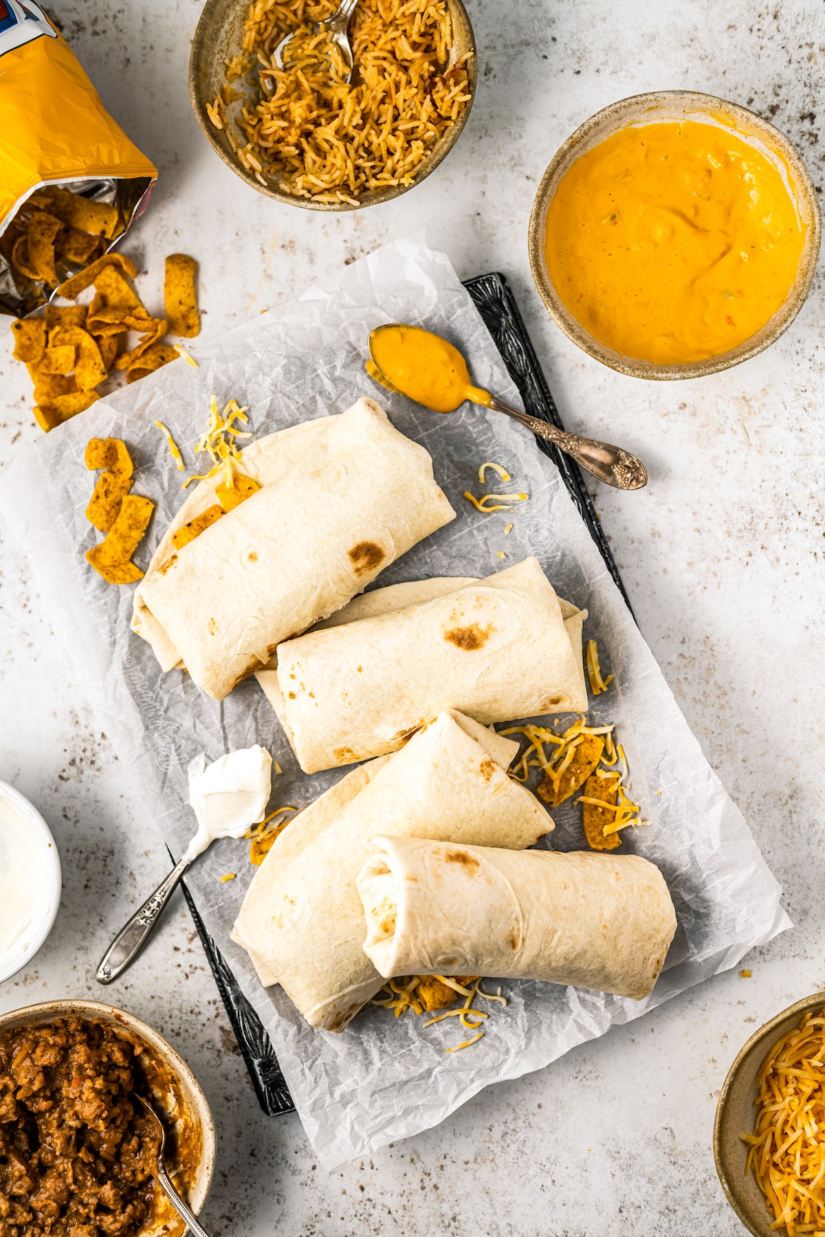 A copycat of the fast-food favorite, this Taco Bell Beefy Melt Burrito is stuffed with seasoned beef, refried beans, rice, Fritos, and more! A tray of homemade burritos with bowls of ingredients arranged around the tray.