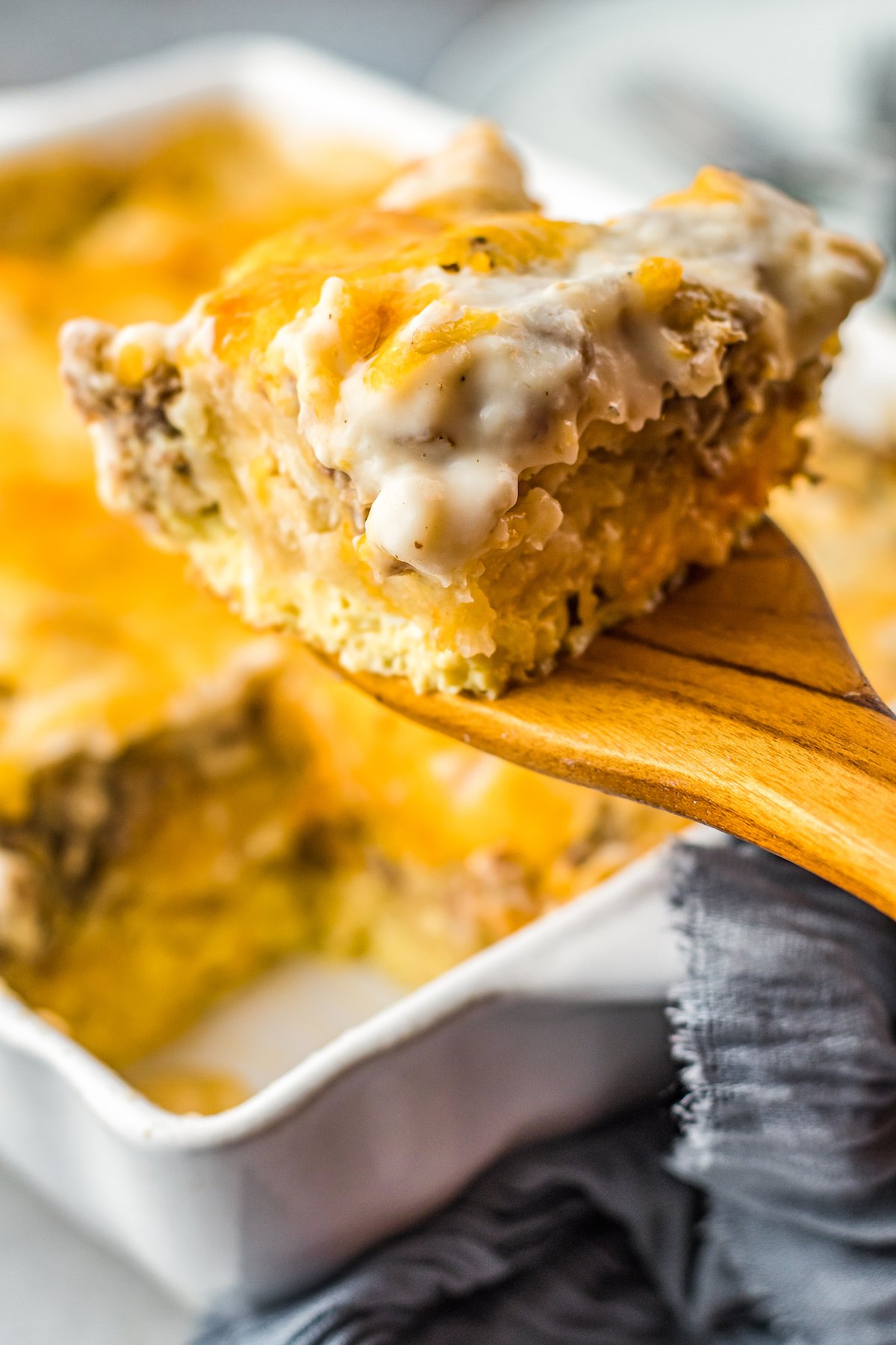 This easy Sausage Biscuits and Gravy Casserole recipe is filled with hearty layers of flaky biscuits, cheesy eggs, and creamy sausage gravy! A serving of biscuits and gravy casserole on a wooden spoon being lifted from the breakfast casserole dish.