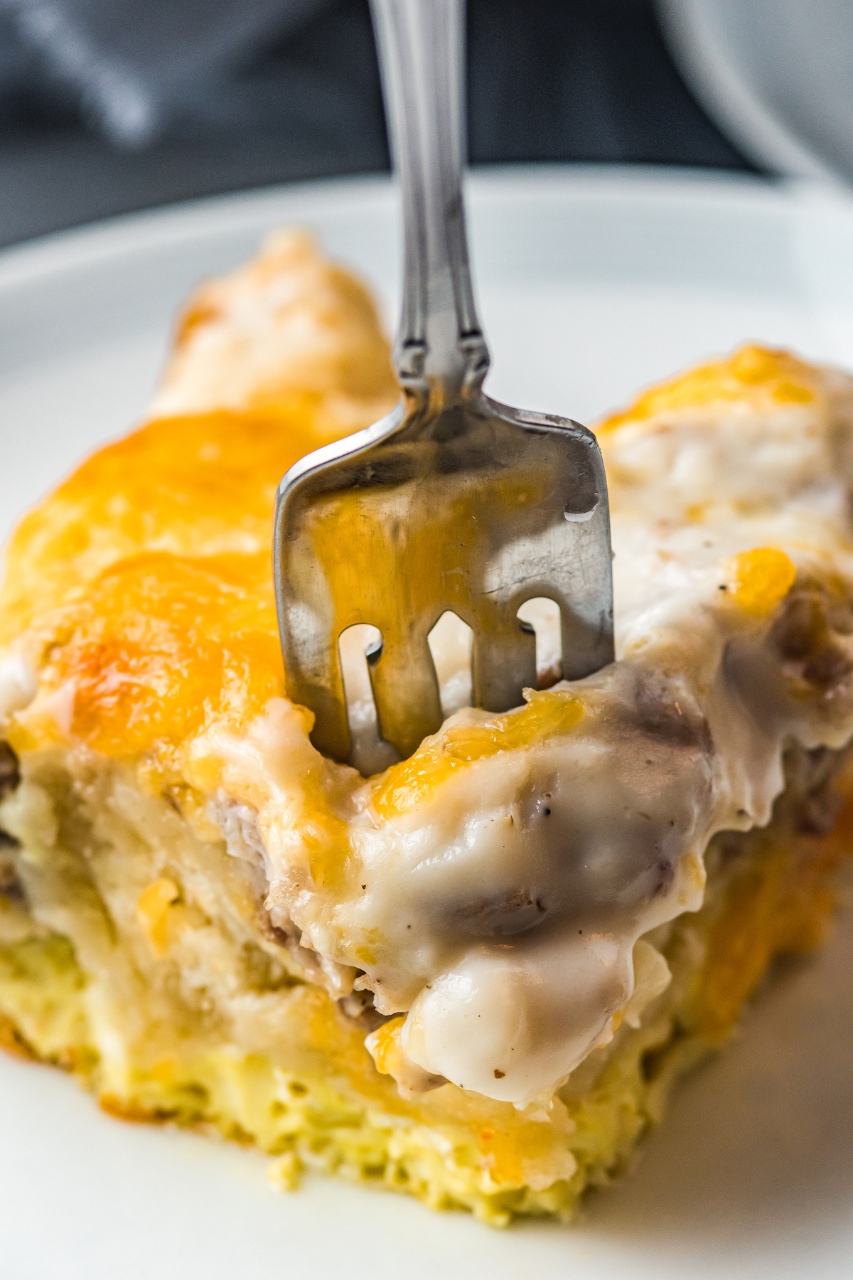 This easy Sausage Biscuits and Gravy Casserole recipe is filled with hearty layers of flaky biscuits, cheesy eggs, and creamy sausage gravy! A serving of creamy, cheesy biscuits and gravy casserole is being pierced with a fork.