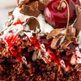 Close-up of a slice of Black Forest poke cake topped with whipped cream, chocolate shavings, cherry sauce, and a whole cherry.