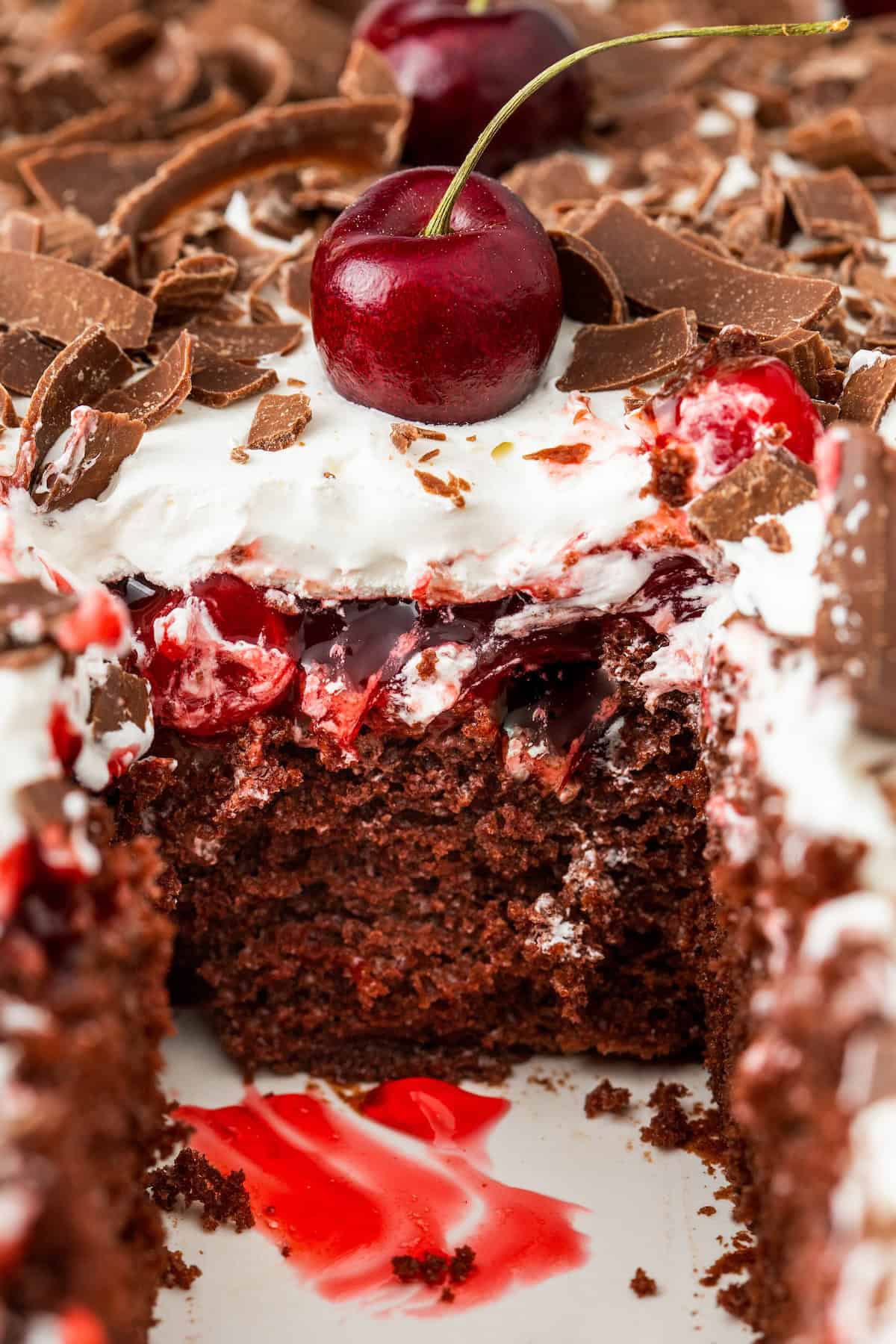 This easy Black Forest Cake recipe is so simple to make with a box of chocolate cake mix, cherry pie filling, and instant chocolate pudding! Close-up of a slice taken from a rich Black Forest poke cake topped with chocolate pudding, vibrant red cherry pie filling, whipped cream, chocolate shavings, and real cherries for garnish.
