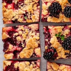 Six blackberry pie bars with crumb topping are arranged on a surface. Some bars are topped with fresh blackberries and a few small green leaves.