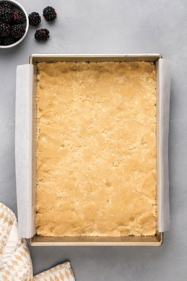 Love pie but hate the hassle? These Blackberry Pie Bars deliver all the flavor of blackberry pie without having to roll out the pie dough! A rectangular baking pan lined with parchment paper contains an unbaked, pressed shortbread crust. Nearby are fresh blackberries in a small bowl and a few scattered on the gray countertop.