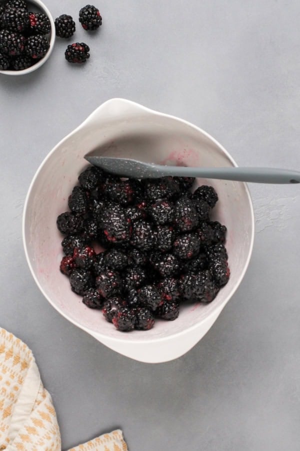 Love pie but hate the hassle? These Blackberry Pie Bars deliver all the flavor of blackberry pie without having to roll out the pie dough! A white mixing bowl filled with blackberries, sugar, cornstarch, and more, being stirred with a gray spatula. With a few blackberries and a small bowl of berries nearby on a gray surface.