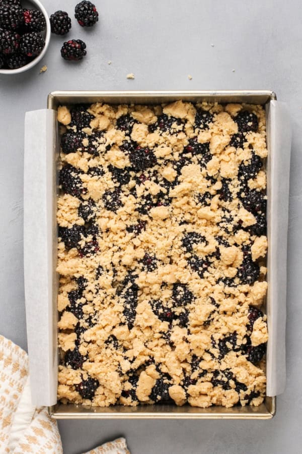Love pie but hate the hassle? These Blackberry Pie Bars deliver all the flavor of blackberry pie without having to roll out the pie dough! A baking pan lined with parchment paper filled with an unbaked blackberry filling topped with crumbly shortbread dough. A bowl of blackberries and a patterned towel are nearby on a gray surface.