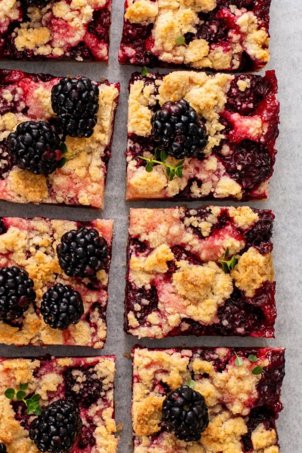 Love pie but hate the hassle? These Blackberry Pie Bars deliver all the flavor of blackberry pie without having to roll out the pie dough! Nine square blackberry crumble bars topped with fresh blackberries and sprigs of green herbs are arranged on a sheet of parchment paper. The bars have a golden, crumbly topping and vibrant berry filling.