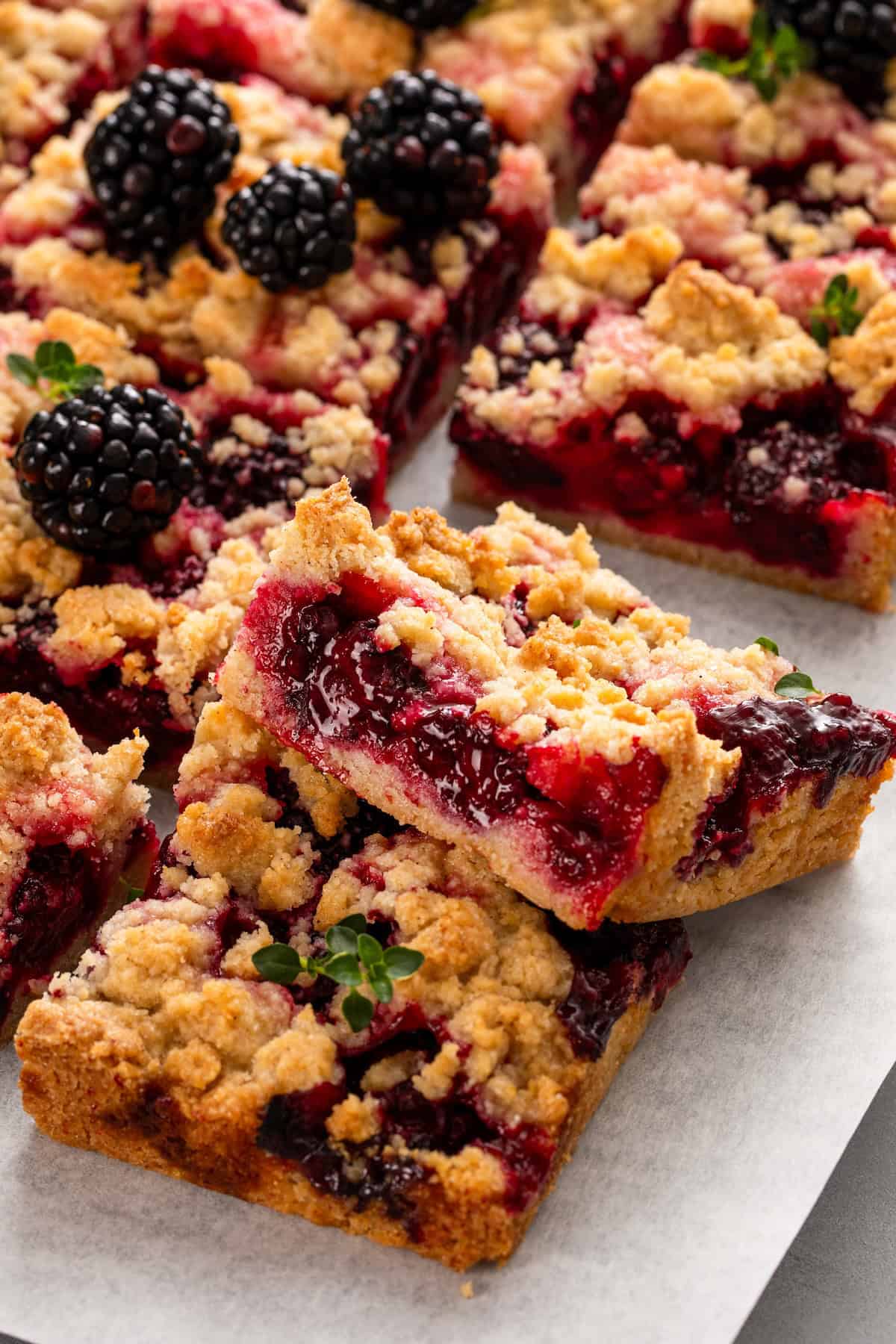 Love pie but hate the hassle? These Blackberry Pie Bars deliver all the flavor of blackberry pie without having to roll out the pie dough! Blackberry pie bars with a golden, crumbly topping are stacked on parchment paper. Fresh blackberries and small green leaves garnish the bars, showing a vibrant, juicy berry filling.
