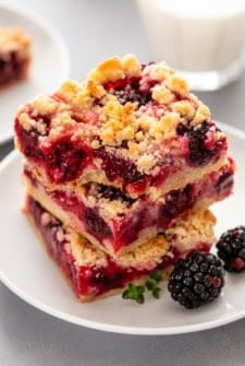 Two crumbly blackberry pie bars stacked on a white plate, filled with blackberries and a golden oat topping, with fresh blackberries and a sprig of thyme next to them. A glass of milk is blurred in the background.