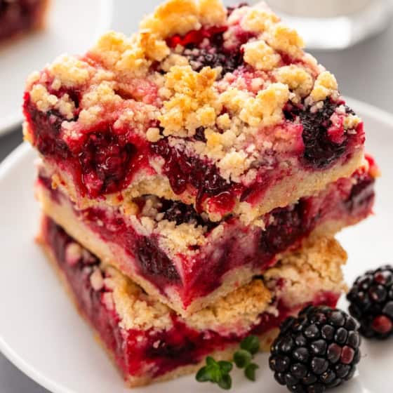 Two crumbly blackberry pie bars stacked on a white plate, filled with blackberries and a golden oat topping, with fresh blackberries and a sprig of thyme next to them. A glass of milk is blurred in the background.