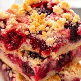 Two crumbly blackberry pie bars stacked on a plate, showing layers of golden crust and juicy blackberry filling. A fresh blackberry and mint garnish are on the plate.