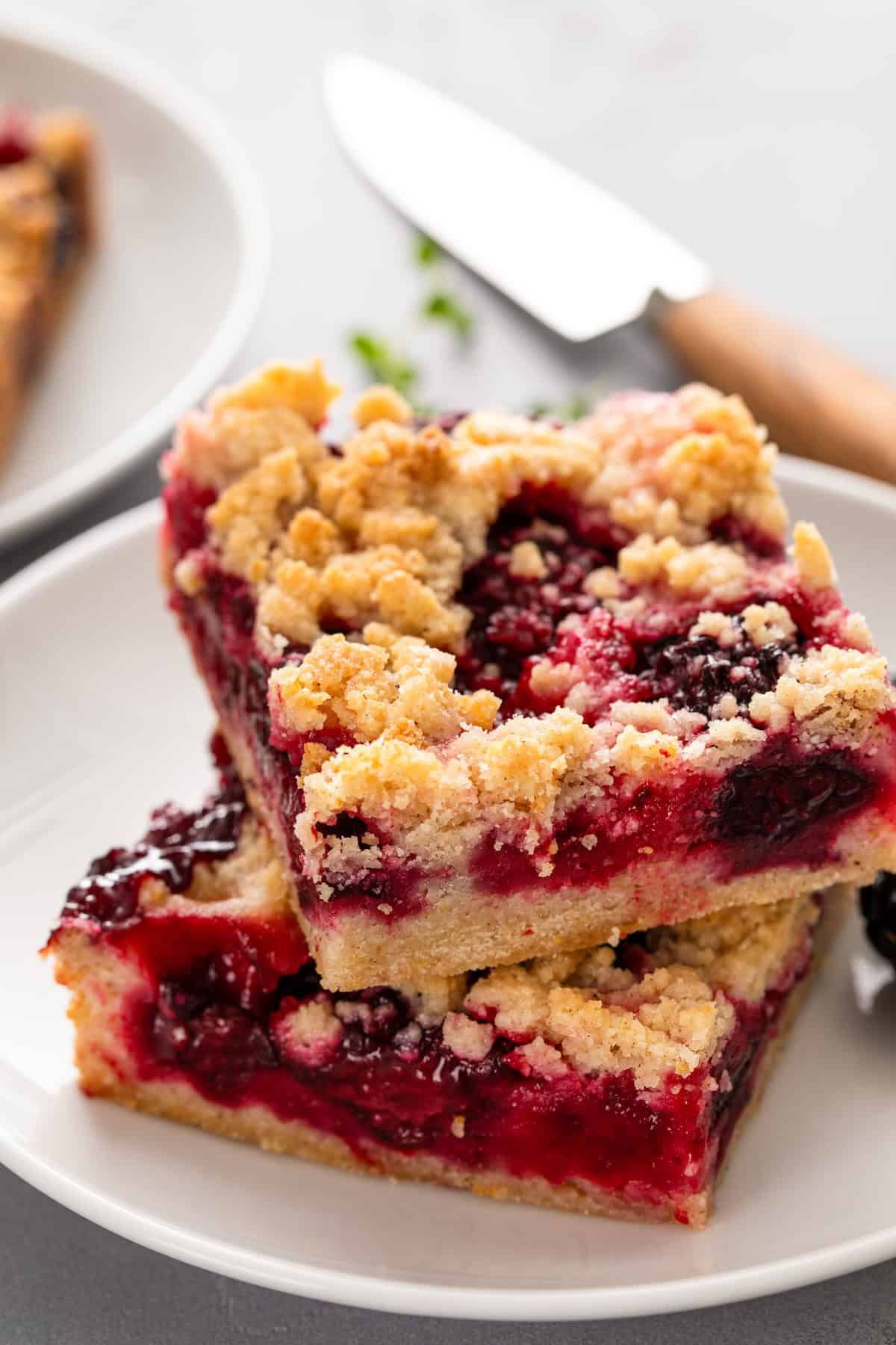 Love pie but hate the hassle? These Blackberry Pie Bars deliver all the flavor of blackberry pie without having to roll out the pie dough! Two stacked slices of blackberry pie bars with a golden, crumbly topping and vibrant red berry filling on a white plate, with a knife and another plate in the blurred background.