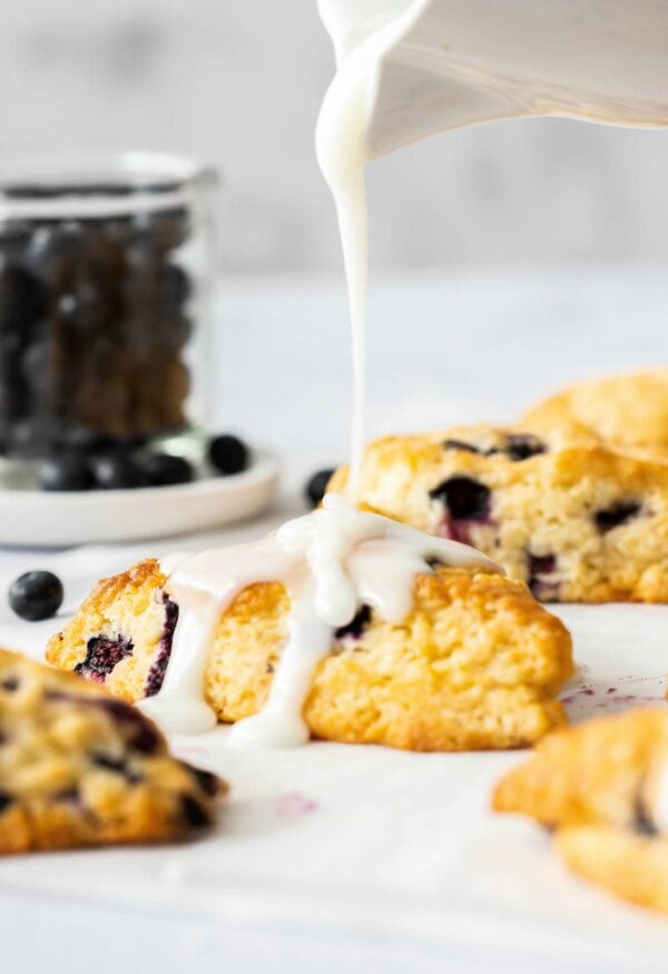 These Blueberry Scones are soft, flaky and bursting with blueberries. Add a creamy vanilla glaze and this easy scone recipe is a real treat! Vanilla glaze being poured over blueberry scones.