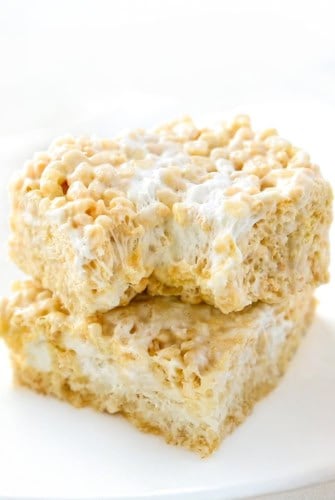 Two gooey Brown Butter Rice Krispie Treats stacked on a white plate, showing crispy cereal bound together with melted marshmallow, with a bite taken out of the top treat.