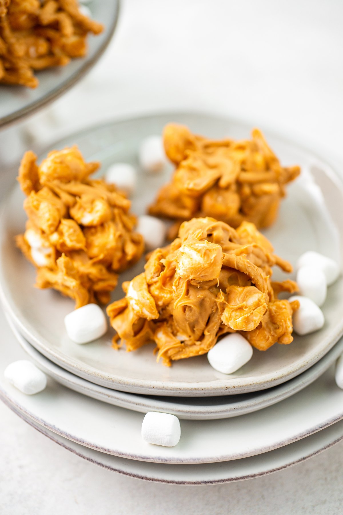 Three no-bake peanut butter haystacks on a plate, with marshmallows scattered over them.
