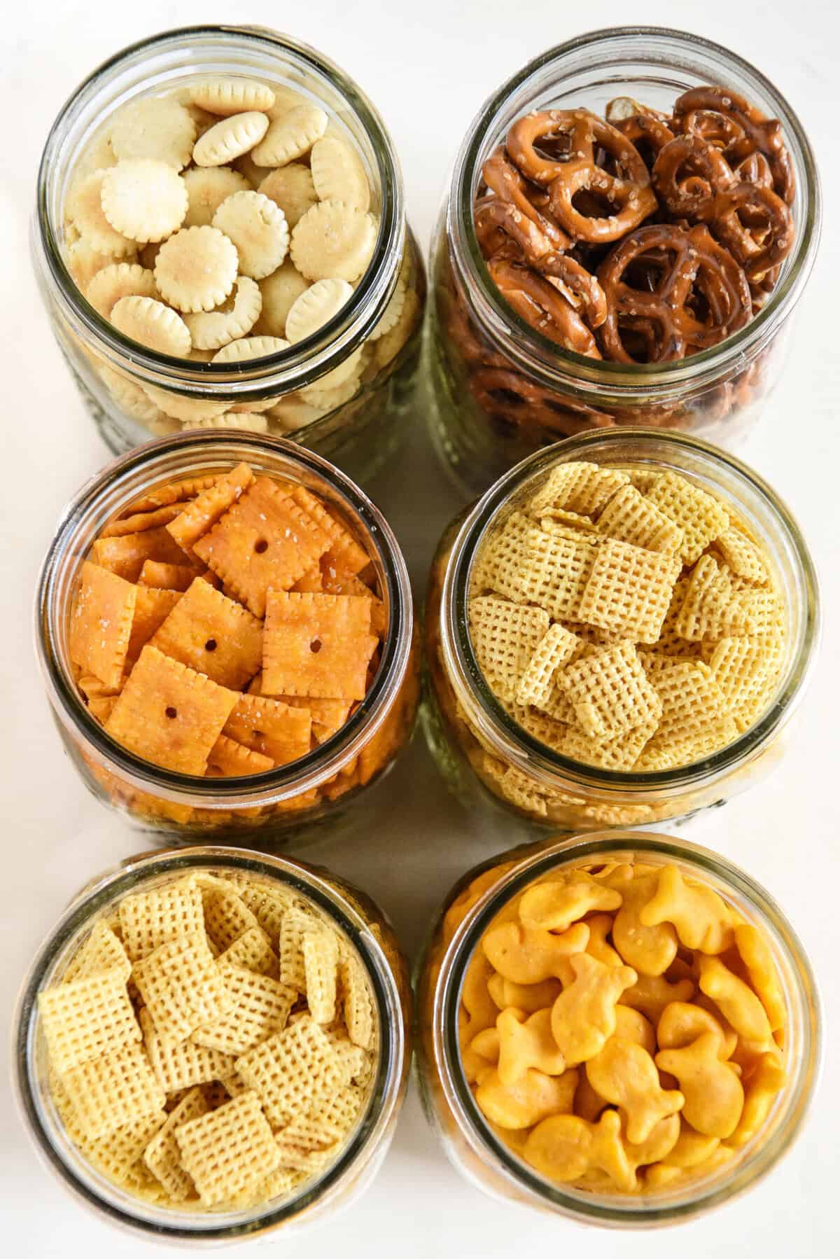 This easy-to-make Homemade Chex Mix recipe is buttery, crunchy, cheesy, slightly spicy, and perfectly savory with a garlicky ranch flavor! Six glass jars filled with different snacks: mini round crackers, pretzels, square cheese crackers, fish-shaped crackers, and two types of Chex cereal, all arranged neatly on a white surface.