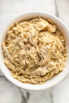 A white bowl filled with creamy shredded crockpot chicken and gravy seasoned with herbs and pepper, served over a bed of rice, on a marble surface.