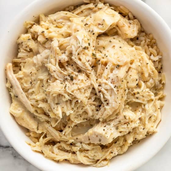 A white bowl filled with creamy shredded crockpot chicken and gravy seasoned with herbs and pepper, served over a bed of rice, on a marble surface.