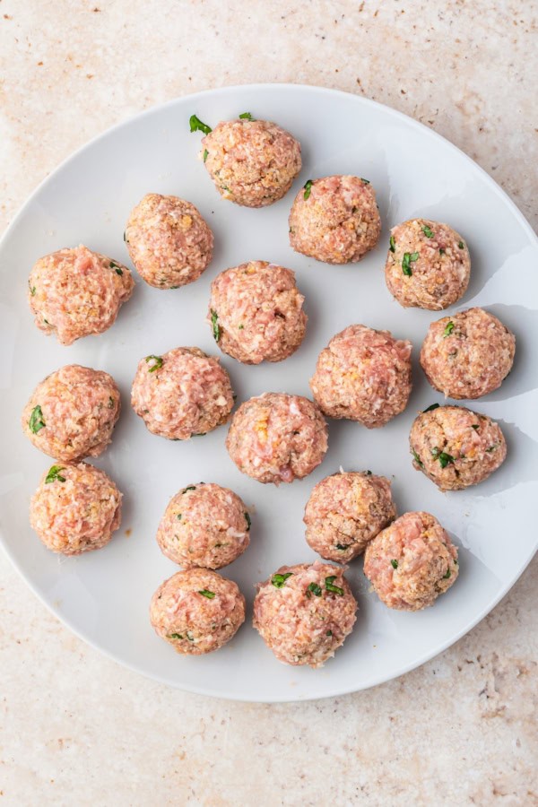 Ready in just 30 minutes, juicy Chicken Piccata Meatballs are simmered in a luscious lemon-caper sauce. Perfect served over pasta or rice! A white plate with several uncooked chicken meatballs, mixed with herbs and breadcrumbs, arranged in a loose circle on a light-colored surface.