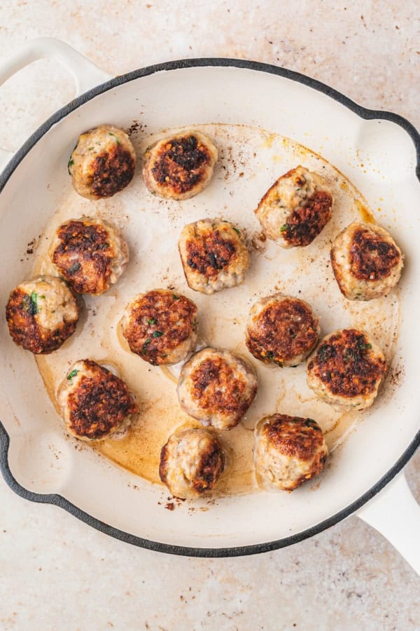 Ready in just 30 minutes, juicy Chicken Piccata Meatballs are simmered in a luscious lemon-caper sauce. Perfect served over pasta or rice! A white skillet with browned chicken meatballs, some with herbs visible, cooking in their juices on a light tan countertop.