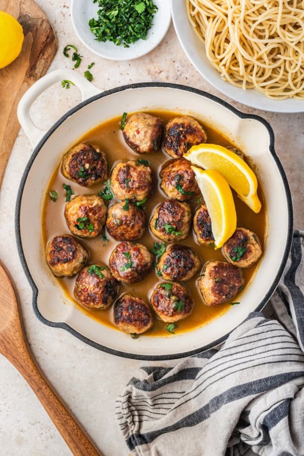 Ready in just 30 minutes, juicy Chicken Piccata Meatballs are simmered in a luscious lemon-caper sauce. Perfect served over pasta or rice! A pan filled with browned chicken meatballs in a lemon-caper sauce, garnished with chopped herbs and lemon wedges, sits next to a bowl of cooked spaghetti, a small plate of herbs, and a striped kitchen towel.