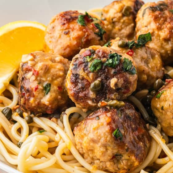 A close-up of spaghetti topped with browned chicken piccata meatballs in a lemon caper sauce, garnished with chopped herbs, and served with lemon wedges in a white bowl.