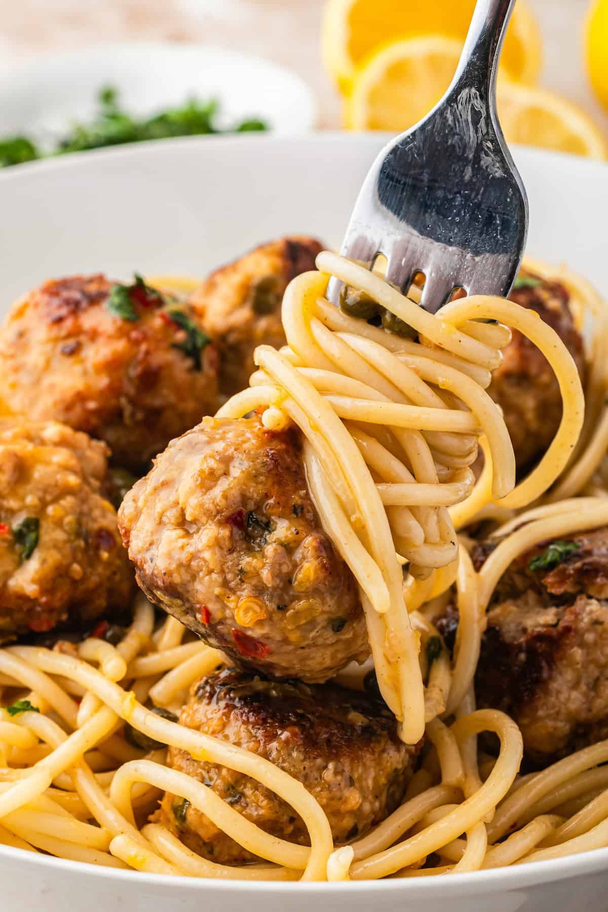 Ready in just 30 minutes, juicy Chicken Piccata Meatballs are simmered in a luscious lemon-caper sauce. Perfect served over pasta or rice! A fork twirls spaghetti noodles above a bowl filled with spaghetti and several chicken piccata meatballs, garnished with fresh herbs. In the background, there are slices of lemon and a plate with chopped parsley.
