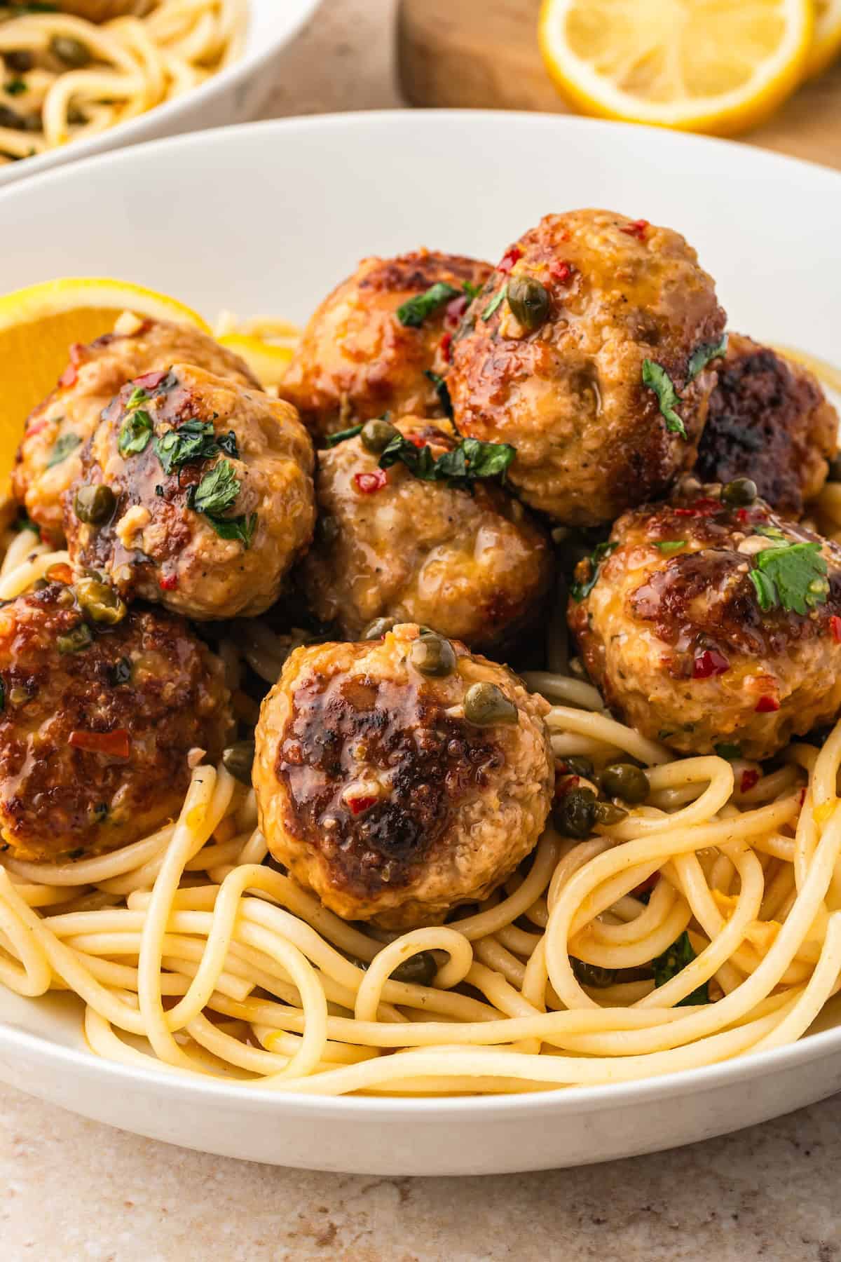 Ready in just 30 minutes, juicy Chicken Piccata Meatballs are simmered in a luscious lemon-caper sauce. Perfect served over pasta or rice! A white bowl filled with spaghetti topped with several browned chicken piccata meatballs in a lemon sauce with capers, garnished with chopped herbs and with a lemon wedge on the side.