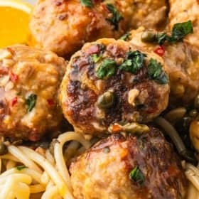Close-up of lemon piccata chicken meatballs garnished with herbs, served on a bed of spaghetti. A slice of lemon is visible in the background, and the dish looks glossy and flavorful.