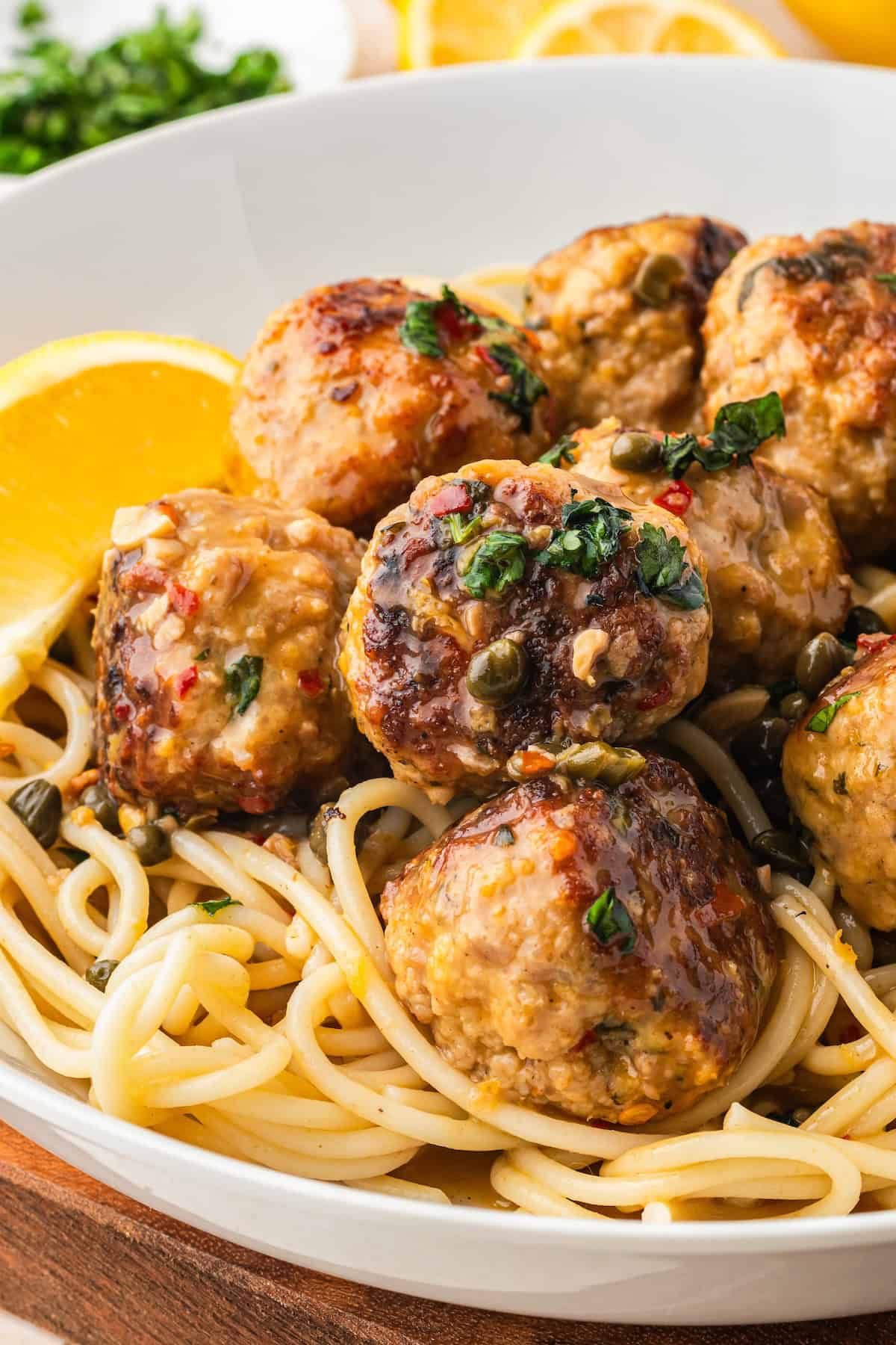 Ready in just 30 minutes, juicy Chicken Piccata Meatballs are simmered in a luscious lemon-caper sauce. Perfect served over pasta or rice! A close-up of spaghetti topped with browned chicken piccata meatballs in a lemon caper sauce, garnished with chopped herbs, and served with lemon wedges in a white bowl.