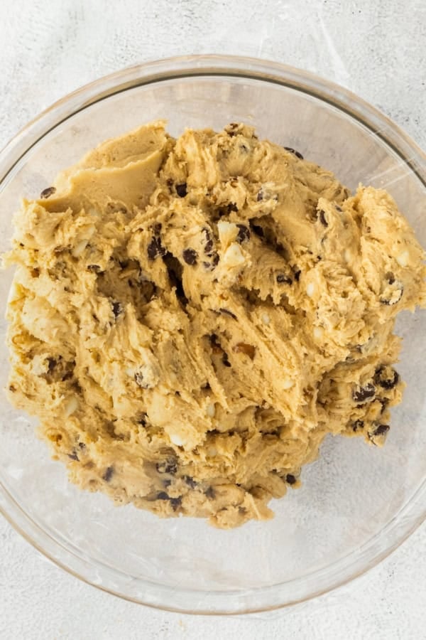 These soft and chewy Chocolate Chip Walnut Cookies have the perfect balance between sweet and nutty with two kinds of chocolate chips. A glass bowl filled with cookie dough sits on a light-colored textured surface. The dough appears creamy and thick, with visible chocolate chips and walnuts mixed throughout.